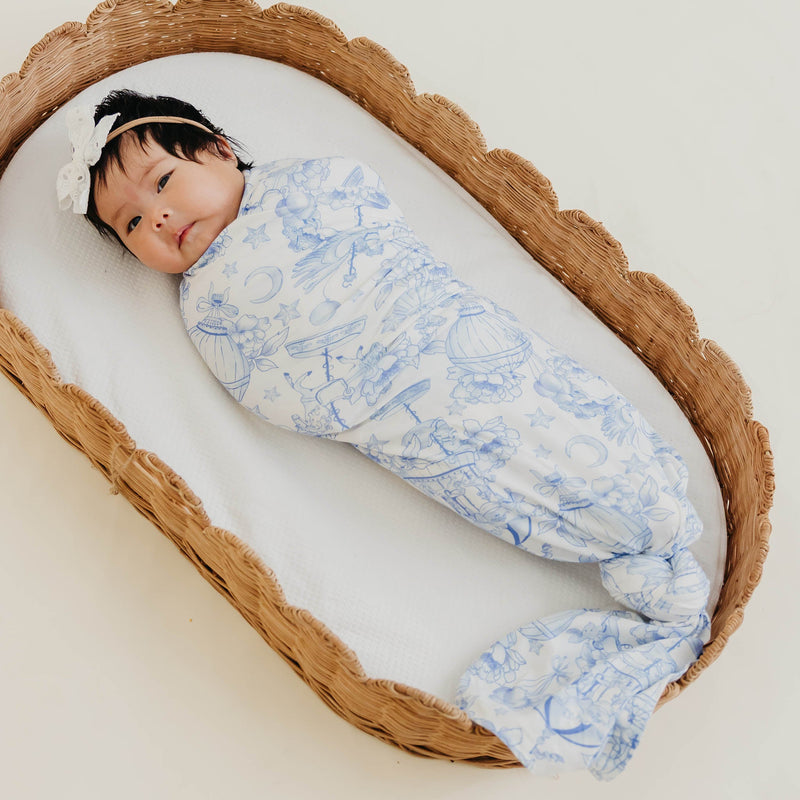 A baby swaddled in a white muslin blanket with a delicate blue toile pattern, lying inside a woven rattan baby bassinet on a light cream background.
