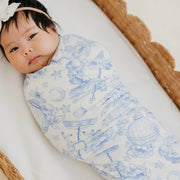 A baby swaddled in a white wrap blanket with a delicate blue celestial print, lying on a white blanket inside a wicker basket. The baby is wearing a beige headband with a white flower.