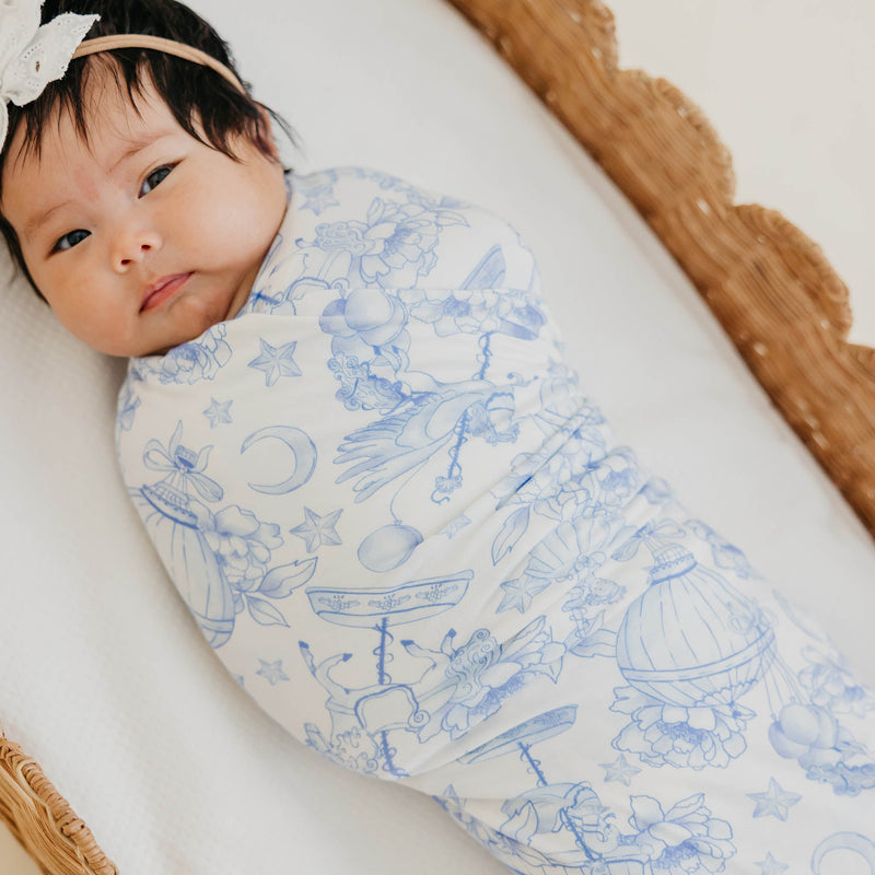 A baby swaddled in a white wrap blanket with a delicate blue celestial print, lying on a white blanket inside a wicker basket. The baby is wearing a beige headband with a white flower.