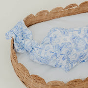 A white knotted baby bonnet with a blue toile-style floral and basket pattern, resting inside a woven wicker basket on a white background.