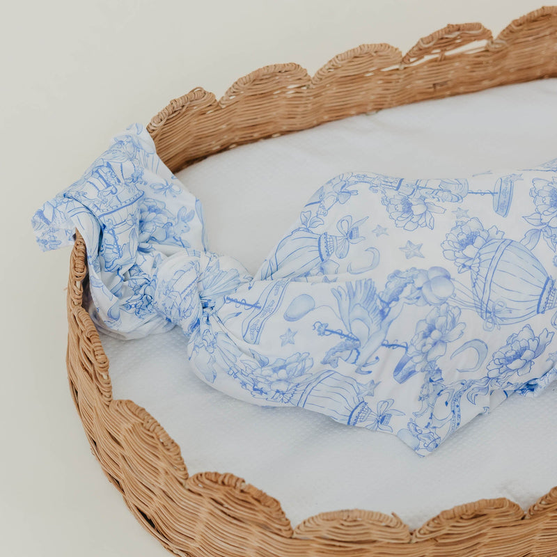 A white knotted baby bonnet with a blue toile-style floral and basket pattern, resting inside a woven wicker basket on a white background.