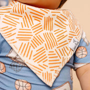 A close-up of a baby wearing a light blue short-sleeve shirt with a basketball print and a cream-colored bandana bib featuring an orange geometric line pattern. A small white tag on the bib reads 'Copper pearl'.