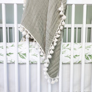 Gray blanket with white pom-poms draped over a crib with green leaf-patterned bedding.