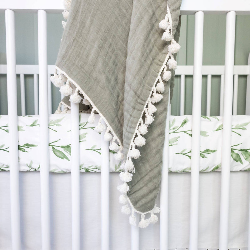 Gray blanket with white pom-poms draped over a crib with green leaf-patterned bedding.