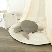 Gray turtle plush toy on a round beige mat with a white curtain in the background