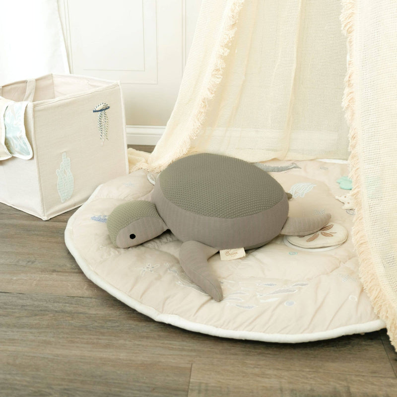 Gray turtle plush toy on a round beige mat with a white curtain in the background