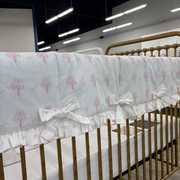 A close-up of a white crib bumper with a pink tree pattern and ruffled trim, draped over a gold metal crib in a showroom.