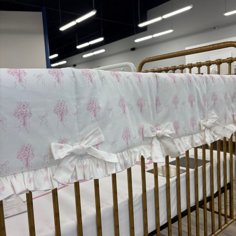 A close-up of a white crib bumper with a pink tree pattern and ruffled trim, draped over a gold metal crib in a showroom.