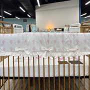 A white crib bedding set with a pink tree pattern and ruffled trim, displayed inside a gold metal crib in a retail showroom.