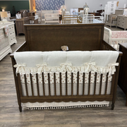 A dark brown wooden crib with a cream-colored bedding set, including a ruffled bumper and a matching skirt, displayed in a furniture showroom.