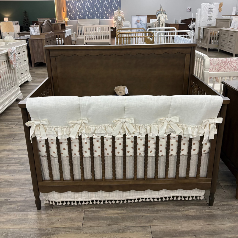 A dark brown wooden crib with a cream-colored bedding set, including a ruffled bumper and a matching skirt, displayed in a furniture showroom.