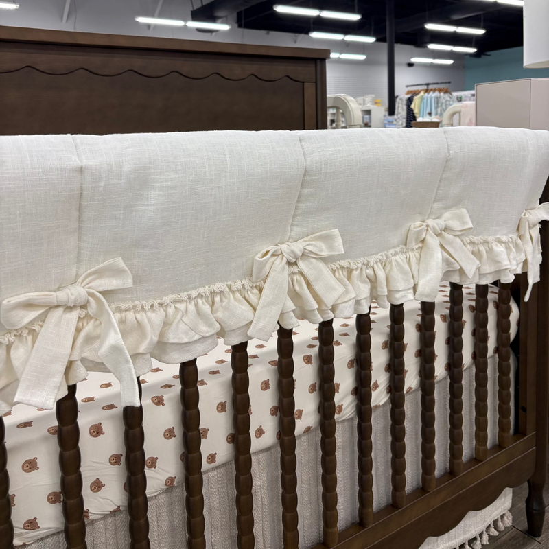A close-up of a nursery crib setup featuring a cream-colored linen crib bumper with ruffled trim and bow details, draped over a dark brown wooden crib. The crib is lined with a matching cream sheet with a small brown pattern.