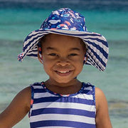 Child wearing a blue and white striped dress and sun hat with ocean design, standing in clear blue water.