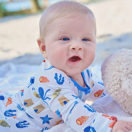 Baby wearing a colorful outfit with sea-themed patterns, lying on a sandy surface.
