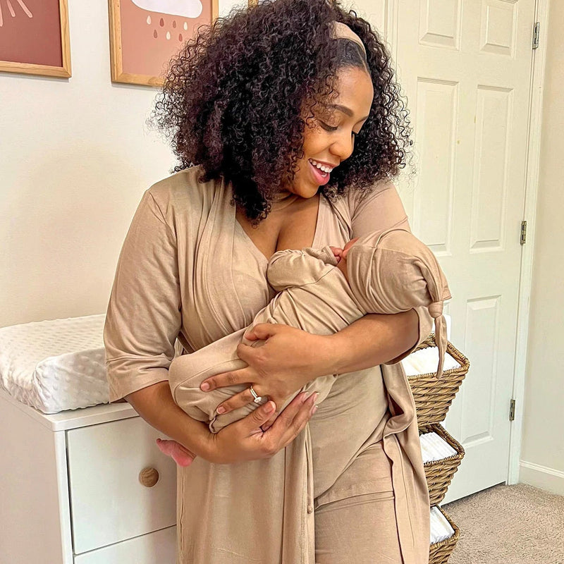 Woman in a beige outfit holding a baby in a sling in a nursery.