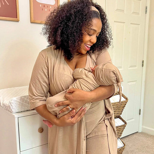 Woman in a beige outfit holding a baby in a sling in a nursery.