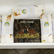 A white wooden display shelf holding several rolled baby blankets or swaddle wraps with deer illustrations and a central printed insert card reading 'Deer Camp.' The setting appears to be a real indoor store or nursery environment with wood flooring visible, so this is Lifestyle rather than Product/Packaging.