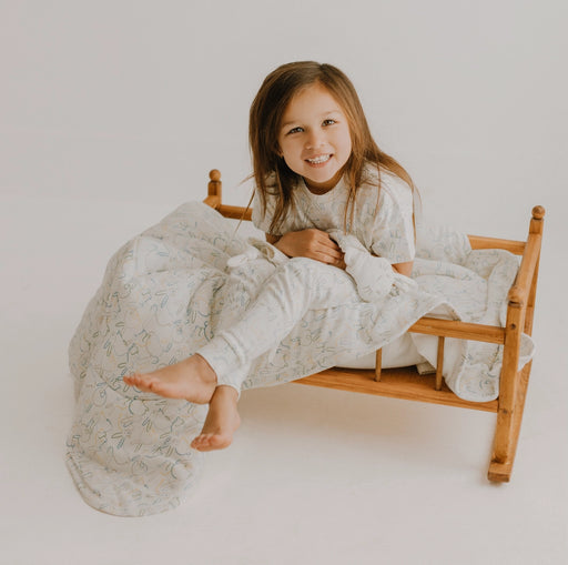 Young girl sitting on a small wooden bed with white bedding against a plain background