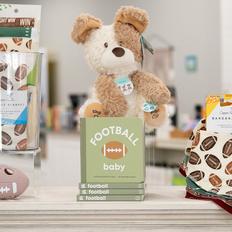 A styled retail display on a white counter featuring a plush brown teddy bear, a green 'FOOTBALL baby' box, a patterned football toy, and other baby items in a softly blurred boutique setting.