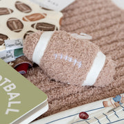 A cozy flat-lay arrangement featuring a plush football-shaped dog toy in taupe and white, resting on a ribbed taupe knit blanket. The scene is styled with a green book, a patterned notebook, and a striped fabric with teddy bear prints.