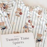 A baby long-sleeve pajama top with a white base, thin navy blue pinstripes, and a teddy bear soccer print, styled flat with a book titled 'Baby's Tummy Time Sports'.