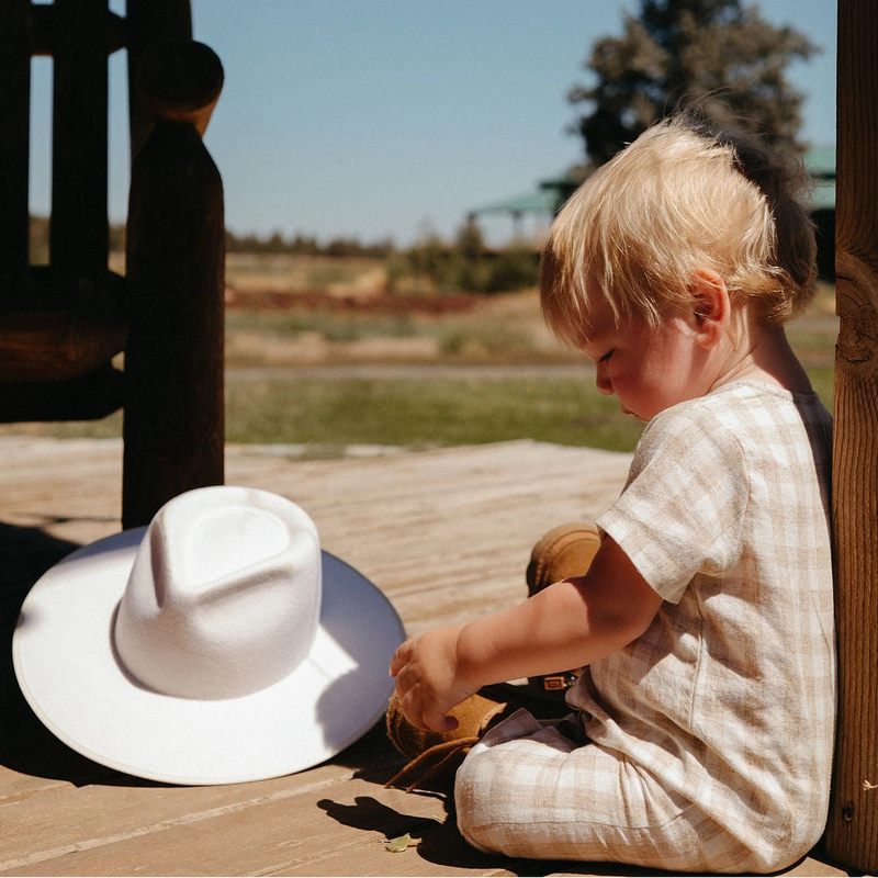 Child sitting on a wooden bench with a white hat nearby, outdoors under a clear blue sky.