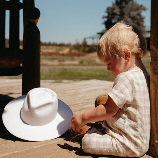 Child sitting on a wooden bench with a white hat nearby, outdoors under a clear blue sky.