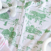 A close-up of a white baby romper or onesie with a green toile-style print of farm animals, trees, and buildings. A white silicone pacifier clip is resting on the garment, and a soft pink fabric is partially visible at the lower left.