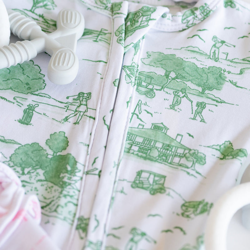 A close-up of a white baby romper or onesie with a green toile-style print of farm animals, trees, and buildings. A white silicone pacifier clip is resting on the garment, and a soft pink fabric is partially visible at the lower left.