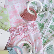 A styled flat-lay of baby clothing and accessories on a white background, including a white long-sleeve romper with pink toile de Jouy print, a green patterned onesie, a pink ruffled fabric piece, and several teething rings and textured toys.