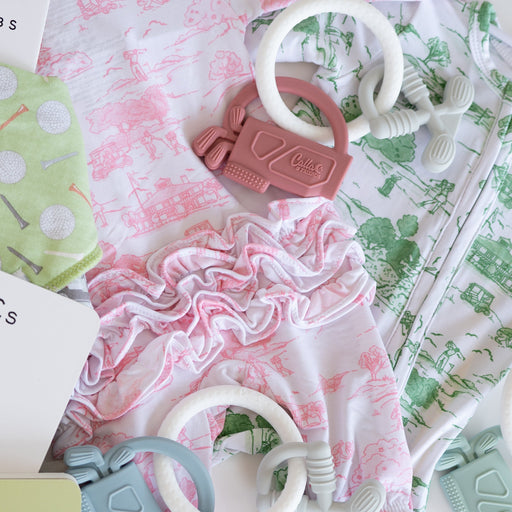 A styled flat-lay of baby clothing and accessories on a white background, including a white long-sleeve romper with pink toile de Jouy print, a green patterned onesie, a pink ruffled fabric piece, and several teething rings and textured toys.