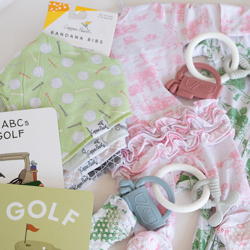 A flat-lay collection of golf-themed baby items, including a green polka dot bandana bib, a pink floral romper, a green and white patterned romper, a baby book titled 'The ABCs of Golf', and a matching golf ball toy.