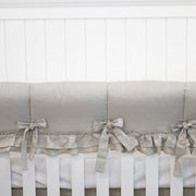 A close-up of a crib bedding set featuring a light beige linen bumper with matching fabric ties and a ruffled trim, shown on a white wooden crib against a white paneled wall.