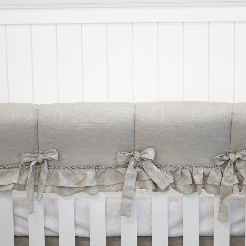 A close-up of a crib bedding set featuring a light beige linen bumper with matching fabric ties and a ruffled trim, shown on a white wooden crib against a white paneled wall.