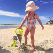 Child in striped swimsuit and sun hat playing on a sandy beach with a shovel and pail.