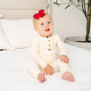 Baby sitting on a bed wearing a white outfit with a red heirloom bow, with a neutral-colored headboard and plant in the background.