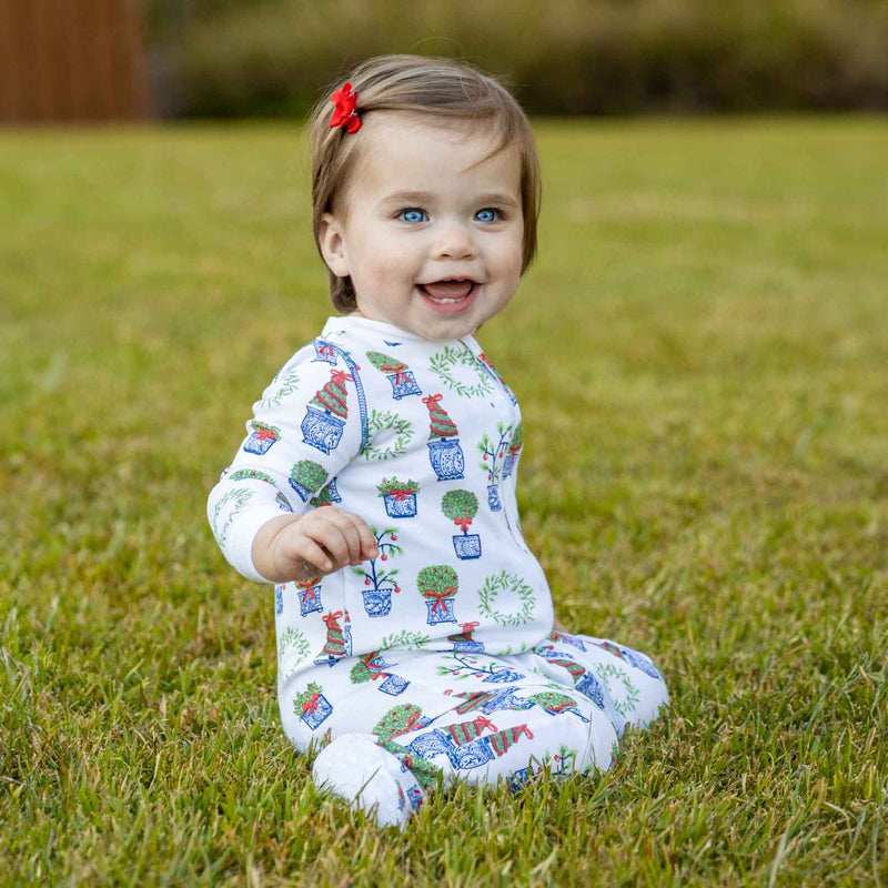 Little blonde girl sitting in the grass wearing Heyward House footed pajama in Christmas Topiaries print