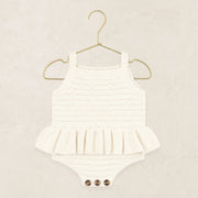 A white ribbed knit baby romper with a ruffled hem and three snap buttons at the crotch, displayed on a gold metal hanger against a textured beige background.