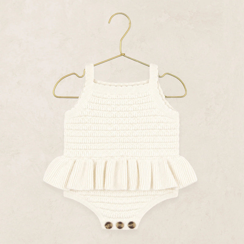 A white ribbed knit baby romper with a ruffled hem and three snap buttons at the crotch, displayed on a gold metal hanger against a textured beige background.