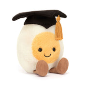 Front view of Jellycat Amuseables Boiled Egg Graduation plush wearing a black mortarboard with gold tassel, seated with corduroy legs