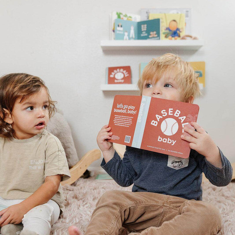 Toddler holding the Baseball Baby board book next to a friend