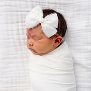 Newborn baby wrapped in white with a large bow headband on a white textured background