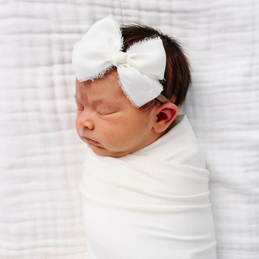 Newborn baby wrapped in white with a large bow headband on a white textured background