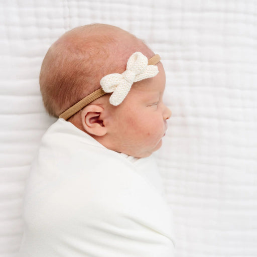 Newborn baby swaddled in white with a decorative headband on a white background