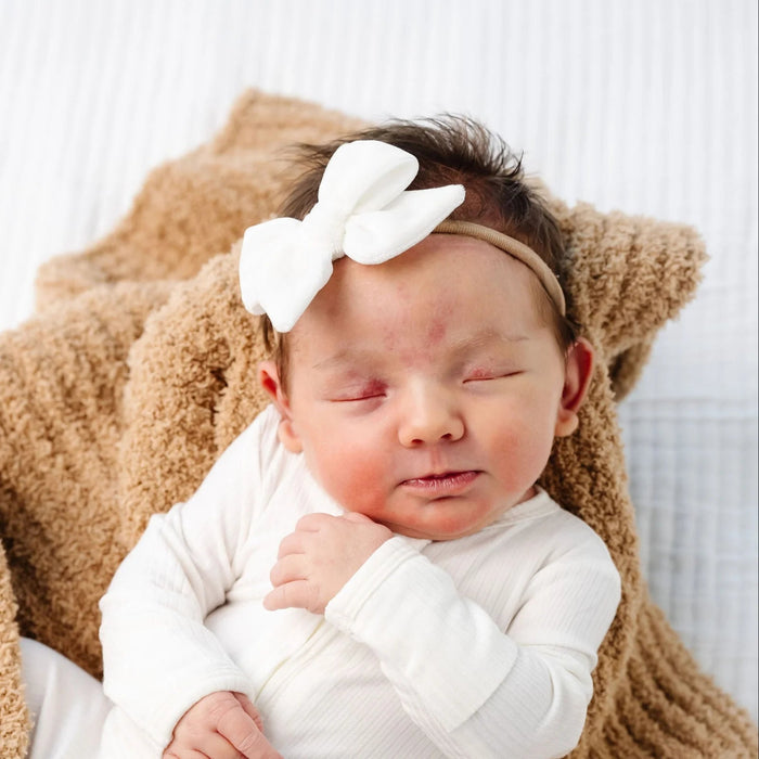 Newborn baby wearing a Lou Lou & Co ivory bow headband with a beige blanket in the background