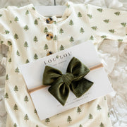Baby onesie with tree pattern and an olive green velvet hair bow on a white fluffy surface