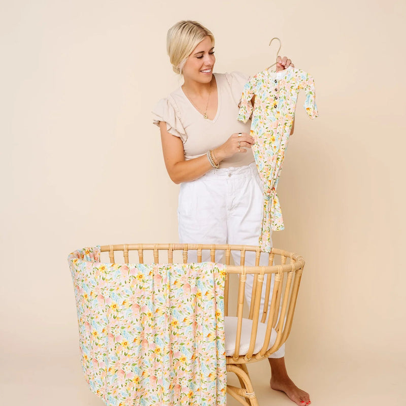A woman stands behind a wooden baby crib holding a matching floral baby romper on a hanger, with a matching floral crib bumper or wrap draped over the front of the crib. The scene is photographed in a studio with a warm beige background.