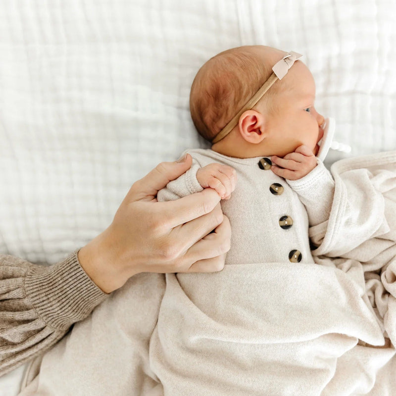 Newborn baby in a beige outfit with a headband, held by an adult hand on a white background