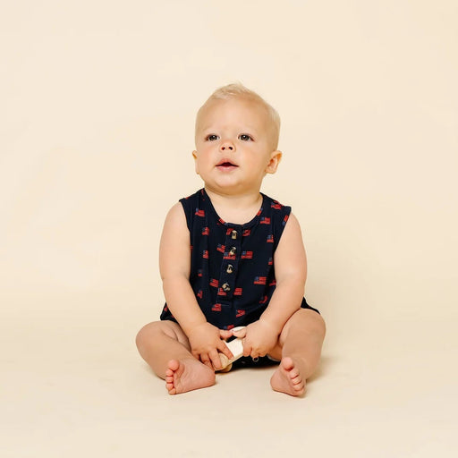 Baby sitting on a beige background wearing a navy sleeveless shirt with red patterns.