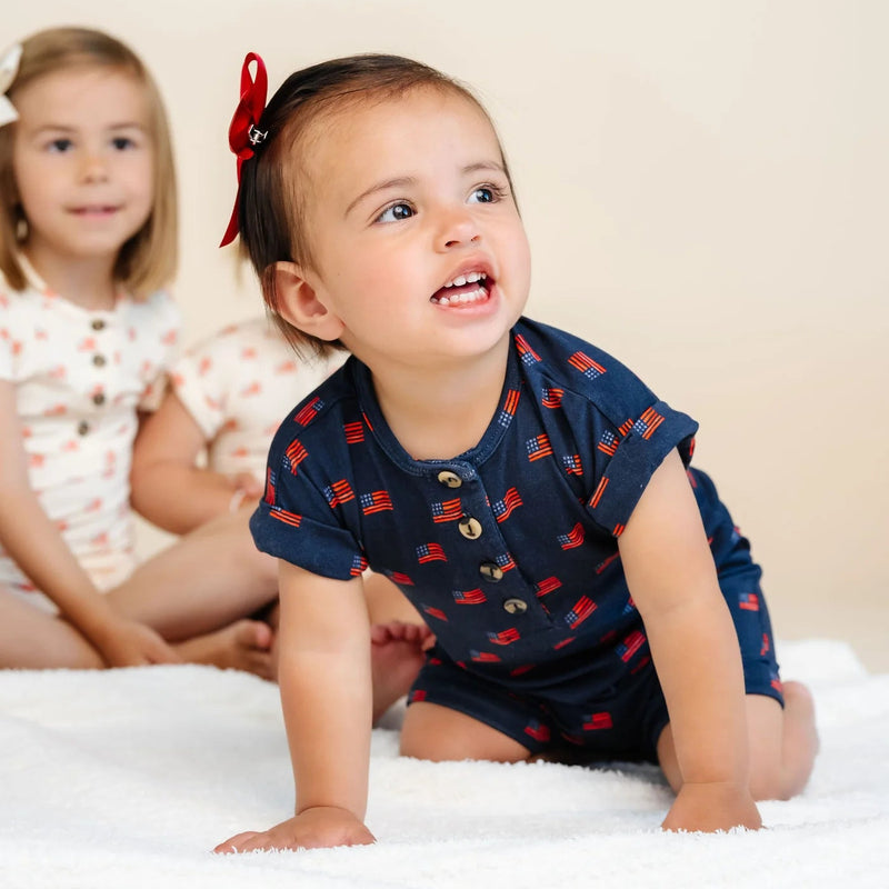 Two children on a bed, one in a navy blue outfit with red patterns and a red bow, the other in a floral dress.